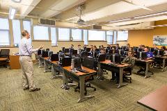 A classroom setting with a teacher presenting to a group of students seated at computer stations.