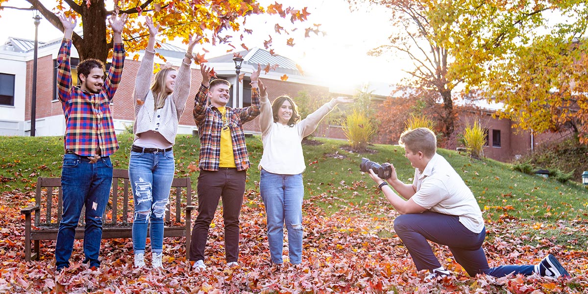Students waving in the air looking happy on a sunny fall afternoon with student photographer taking their photo Students waving in the air looking happy on a sunny fall afternoon with student photographer taking their photo