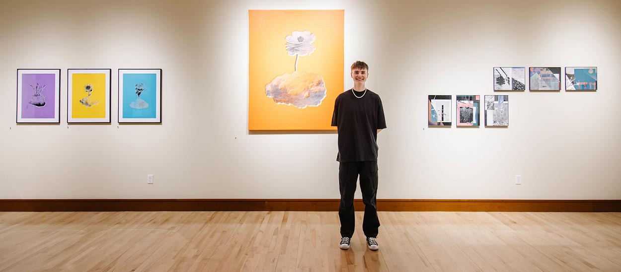 Student smiling standing in a gallery with his protfolio displayed in frames on the wall behind him Student smiling standing in a gallery with his protfolio displayed in frames on the wall behind him