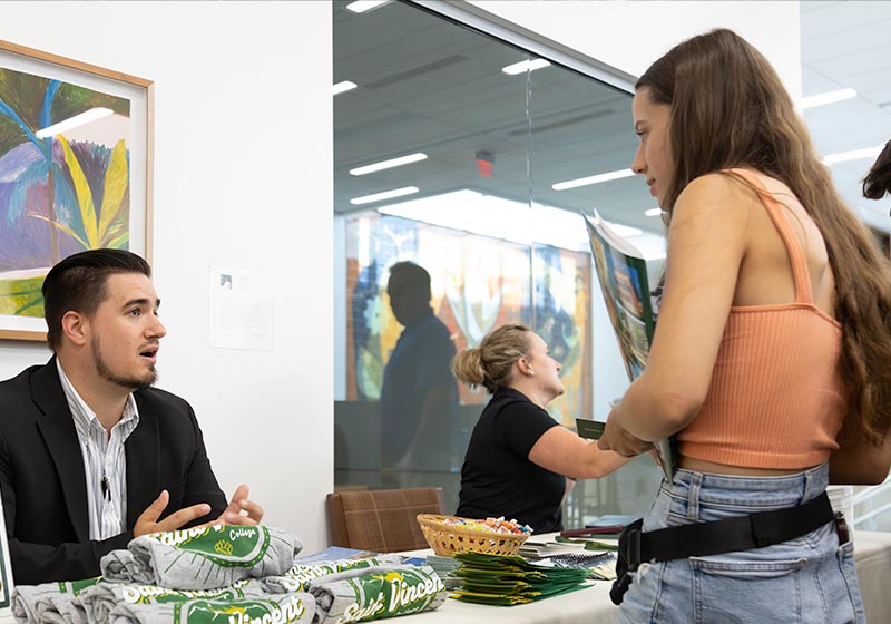 A man in a black suit speaking to a woman in an orange tank top at a registration table, while another woman is seen in the background.