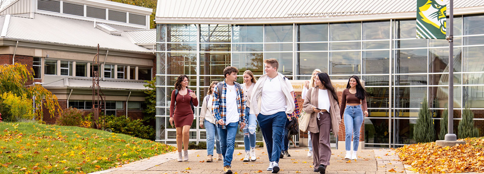 A group of diverse college students walking together outside a modern campus building on a sunny autumn day.