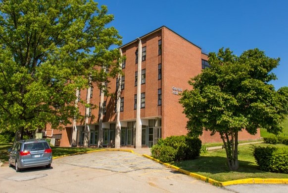 A brick building with large windows surrounded by trees and a parking area.