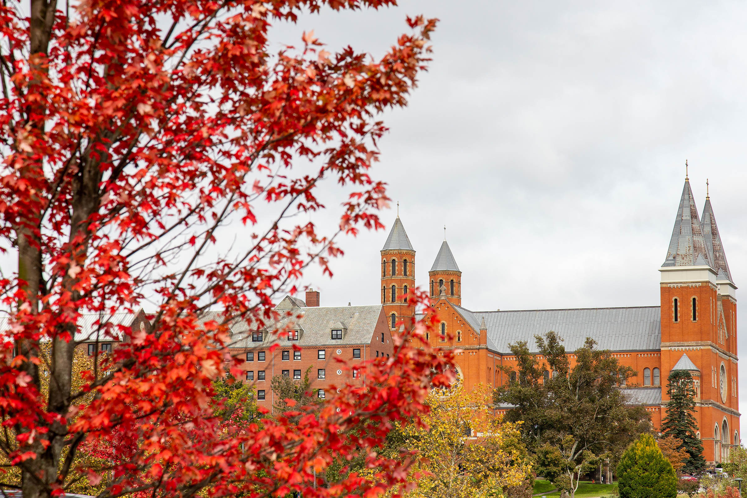 Autumn foliage with vibrant red leaves in the foreground, showcasing a historic brick building with two spires in the background.