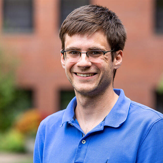Friendly portrait of a man with glasses smiling outdoors, wearing a blue polo shirt.