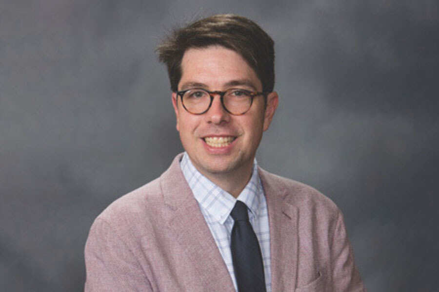 A professional portrait of a man wearing glasses, a light pink blazer, and a blue tie against a blurred background.