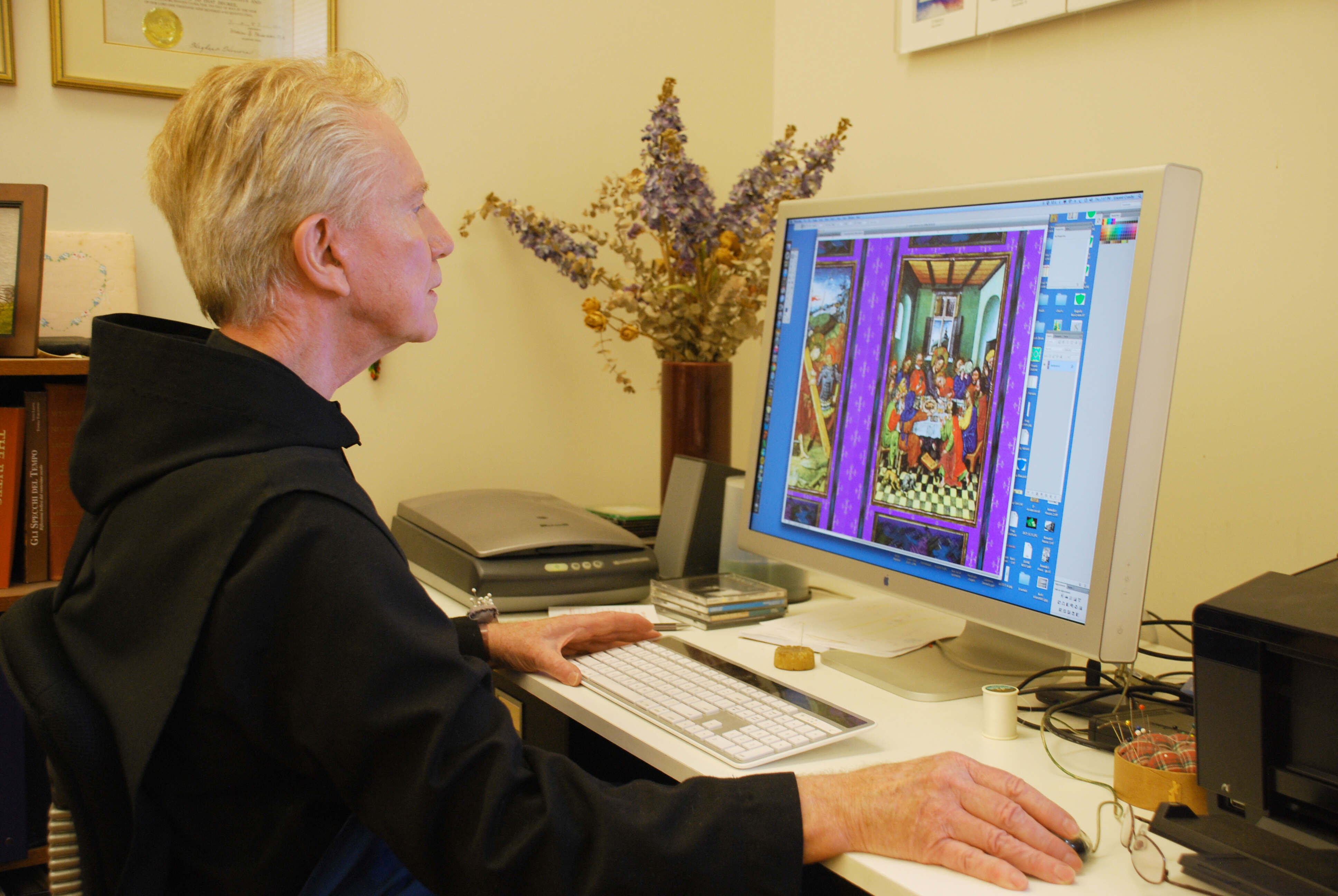 A person focused on editing an image on a computer, surrounded by flowers and office supplies.