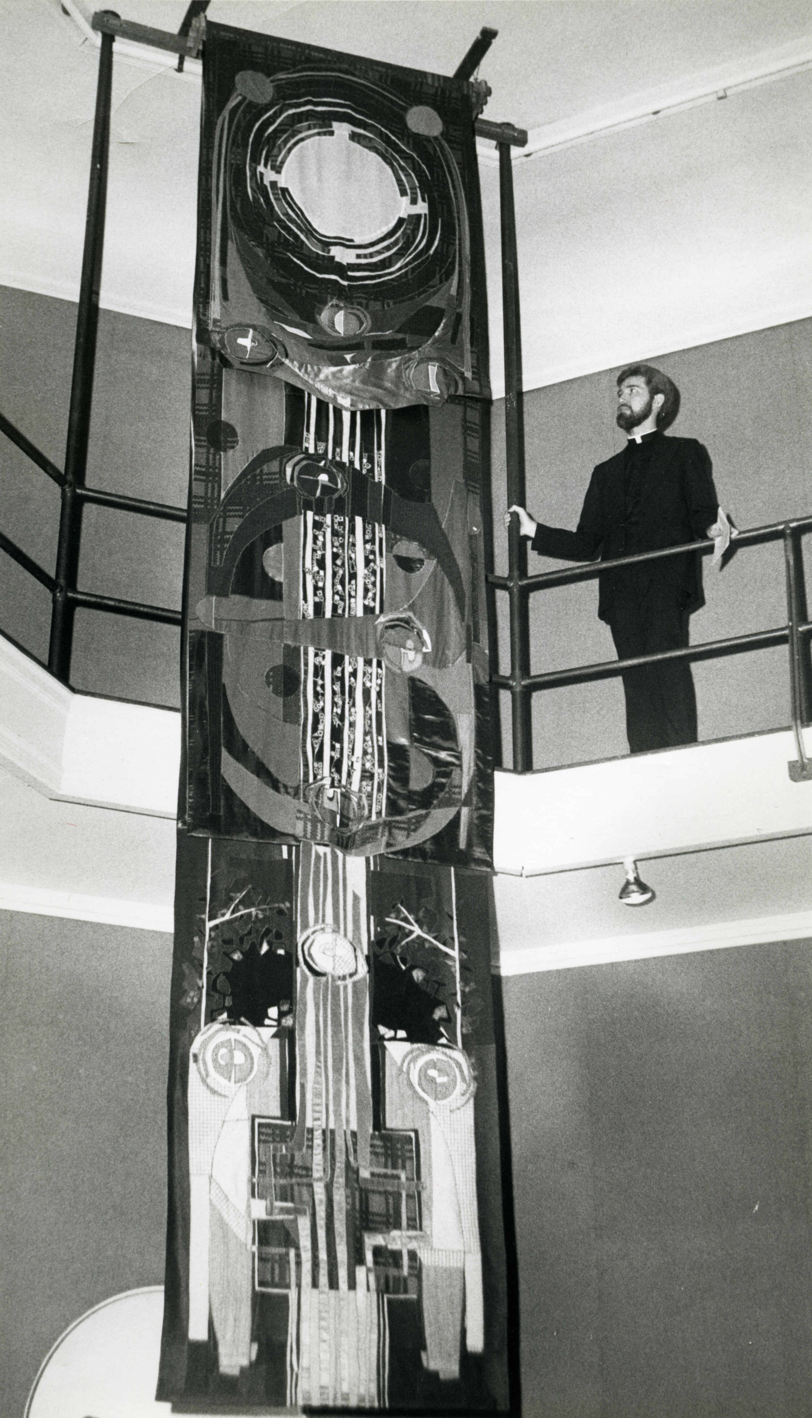 A black and white photograph showcasing a man standing beside a large, abstract tapestry hanging from a balcony.