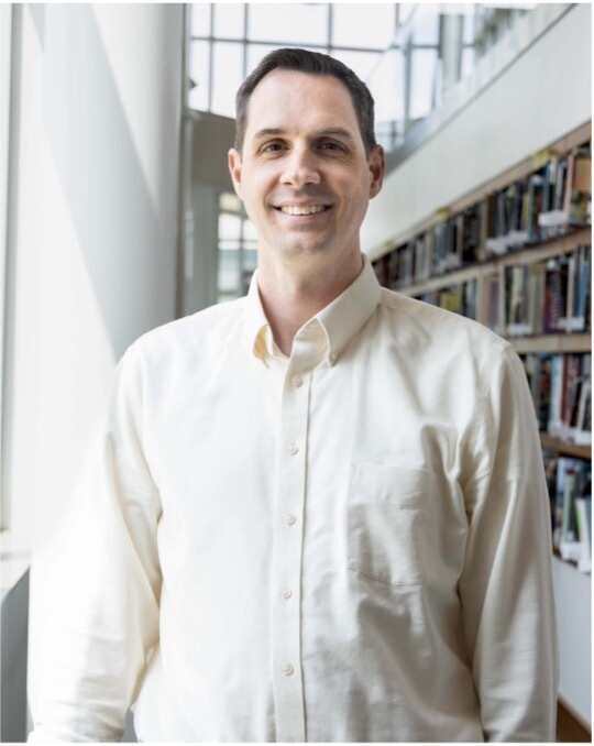 Smiling man in a light-colored shirt standing in a library filled with books.