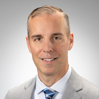 Professional headshot of a man in a light gray suit and blue tie, smiling against a neutral background.