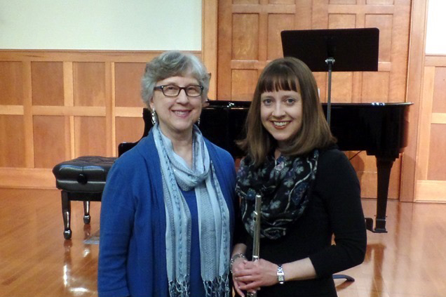 Two musicians pose together in a warm, wooden-paneled music room, with a grand piano in the background.