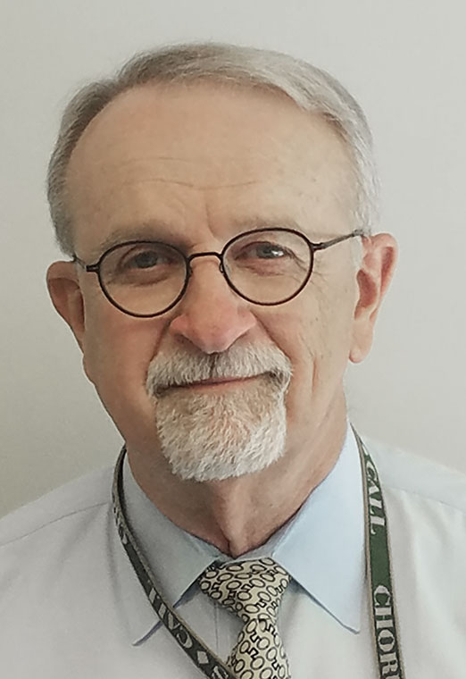 Portrait of a distinguished man with glasses, gray hair, and a goatee, wearing a light blue shirt and a patterned tie.