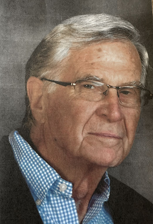 A close-up portrait of an older man with gray hair and glasses, wearing a blue checkered shirt beneath a dark blazer.