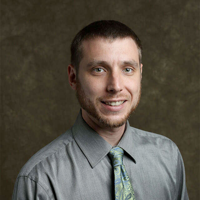 Professional headshot of a man in a gray shirt with a patterned tie, smiling against a textured background.