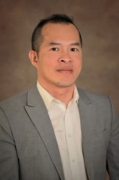 Professional headshot of a man wearing a light gray suit and white shirt, with a neutral expression against a soft, brown background.