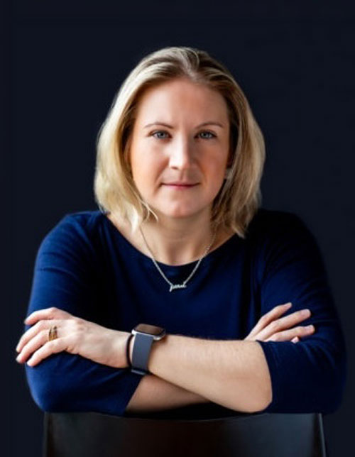 Portrait of a woman with blonde hair in a navy top, arms crossed, against a dark background.