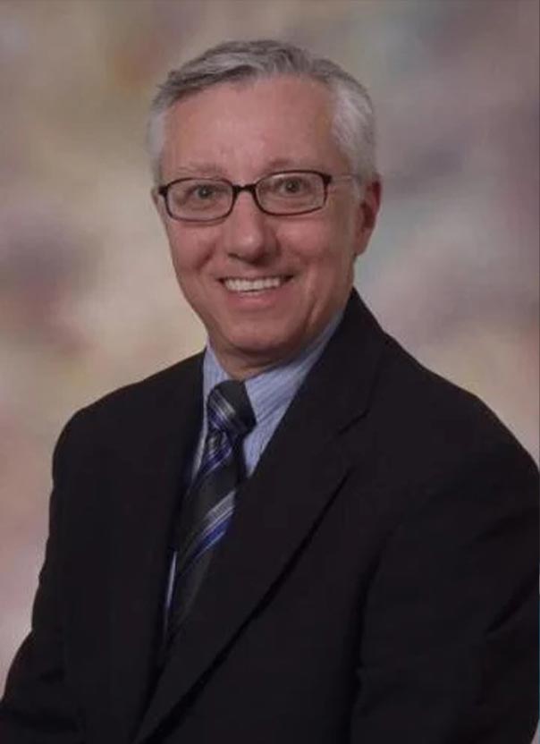 Studio headshot of a smiling man with gray hair and glasses wearing a dark suit against a blurred background.