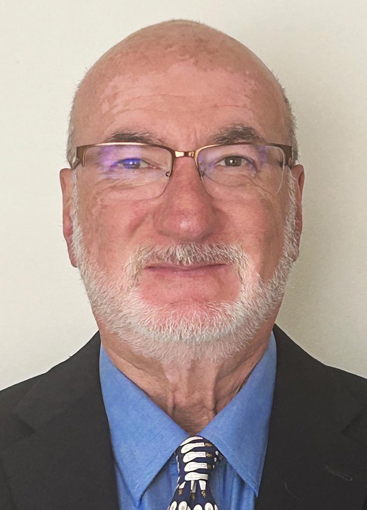 Close-up of an man with glasses and a white beard, wearing a dark suit against a plain background.
