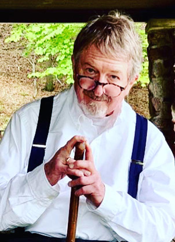 A man with gray hair and glasses, wearing a white shirt and suspenders, holds a wooden cane while sitting outdoors.