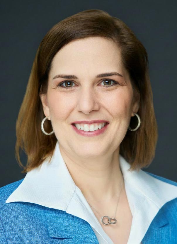 Professional headshot of a smiling woman with shoulder-length brown hair, wearing a blue blazer over a white collared shirt.