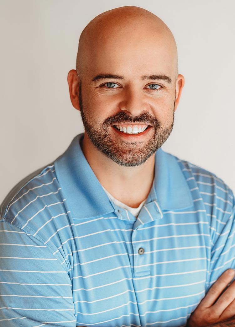 Smiling man with a short beard wearing a light blue striped polo shirt against a neutral background.