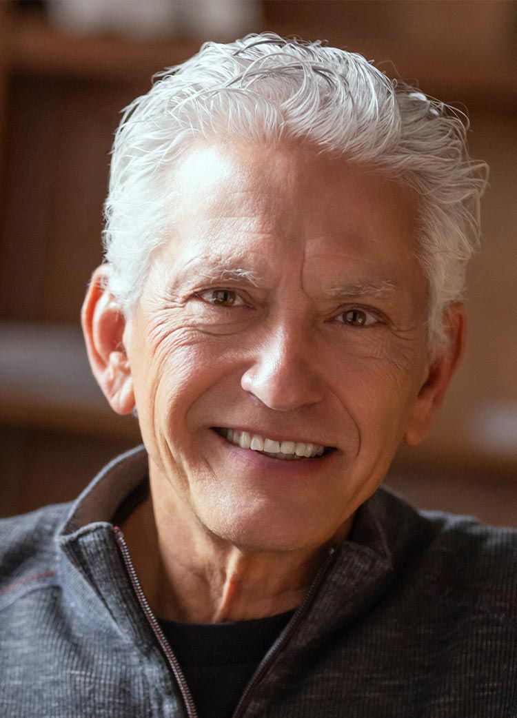 Close-up portrait of a man with short white hair smiling, wearing a gray zip-up sweater in an indoor setting.