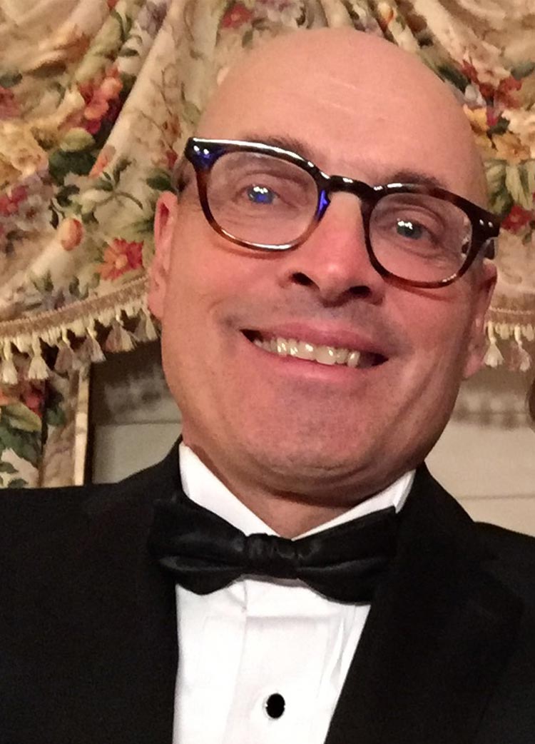 Close-up of a smiling man wearing glasses and a black tuxedo with a bow tie, in front of floral curtains.
