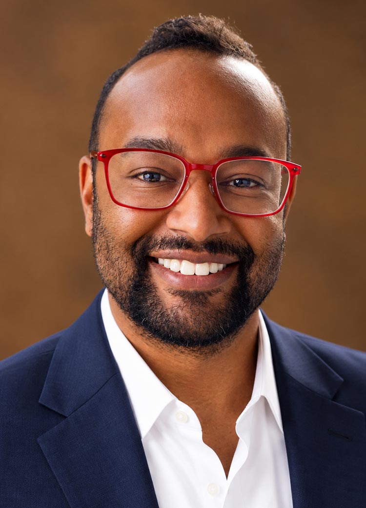 Smiling man wearing red glasses and a navy blazer in a close-up portrait against a brown background.