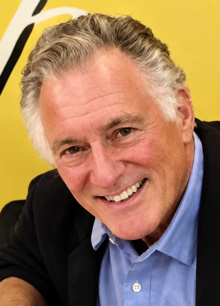 Close-up portrait of a smiling man with gray hair, wearing a blue shirt and dark blazer against a yellow background.
