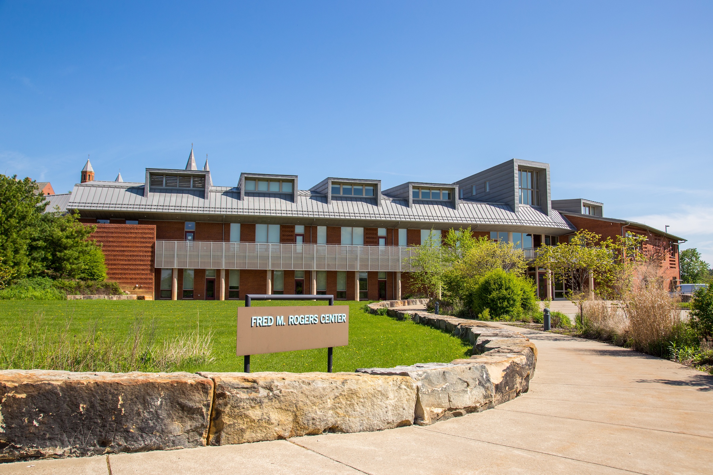Exterior of the Fred M. Rogers Center building with a sign in the foreground and a walkway bordered by landscaping under a clear blue sky.